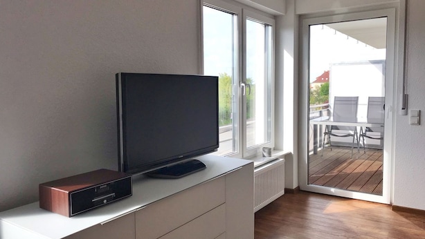 A modern living room with a flat-screen TV on a white cabinet, large windows, and a view of the outside. - Ferienappartement "langschläfer" (Sassnitz)