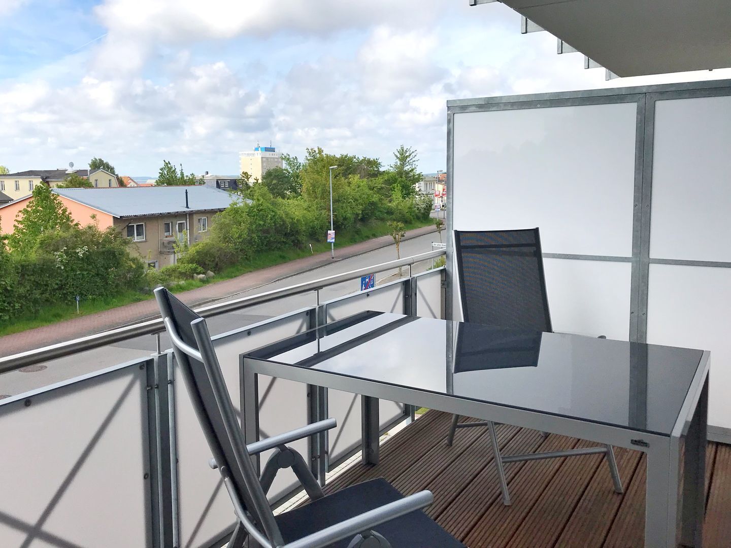 Un balcone con tavolo e sedie, affacciato su una strada con edifici e alberi.