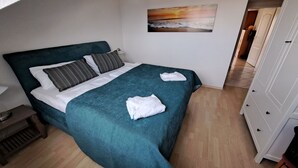 A neatly made bed with a teal bedspread and white linens, a small wooden nightstand with a lamp, and a painting of a sunset on the wall. - Ferienwohnung "osterholz" (Baabe)