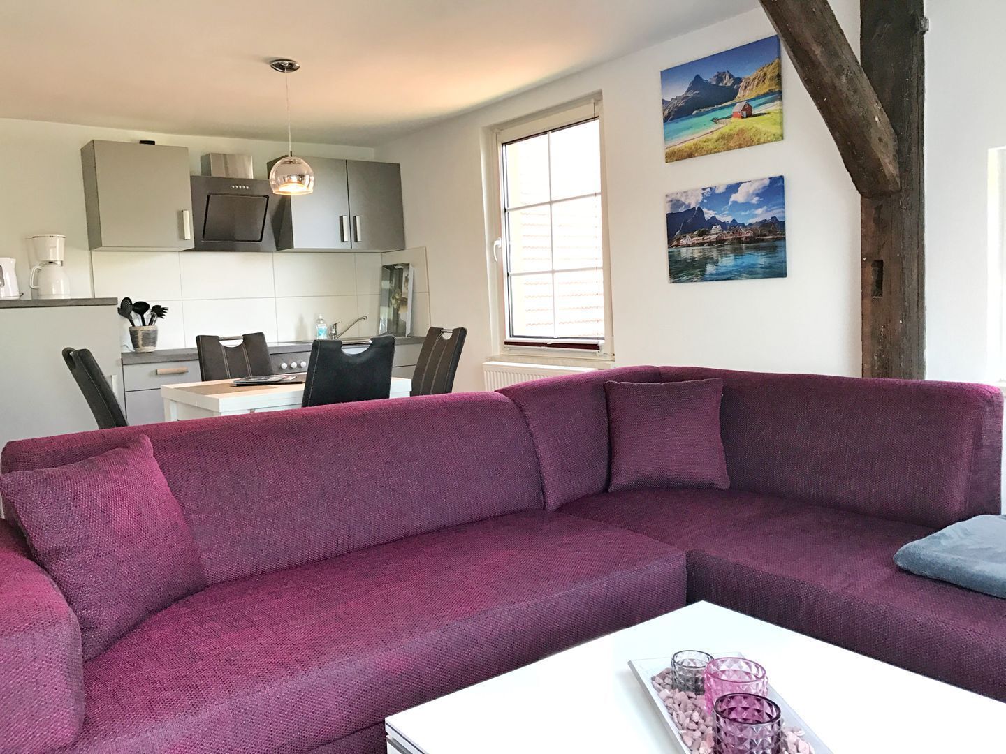 A modern living room with a purple sofa, a dining area with a white table and chairs, and a kitchen with grey cabinets and a hanging light fixture.