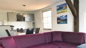 A modern living room with a purple sofa, a dining area with a white table and chairs, and a kitchen with grey cabinets and a hanging light fixture.