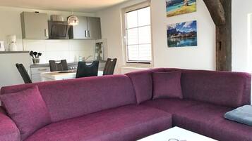 A modern living room with a purple sofa, a dining area with a white table and chairs, and a kitchen with grey cabinets and a hanging light fixture.
