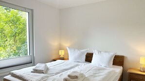 Una cama doble bien hecha con ropa blanca y toallas, un cabecero de madera, lámparas de noche y una ventana con vistas a follaje verde.