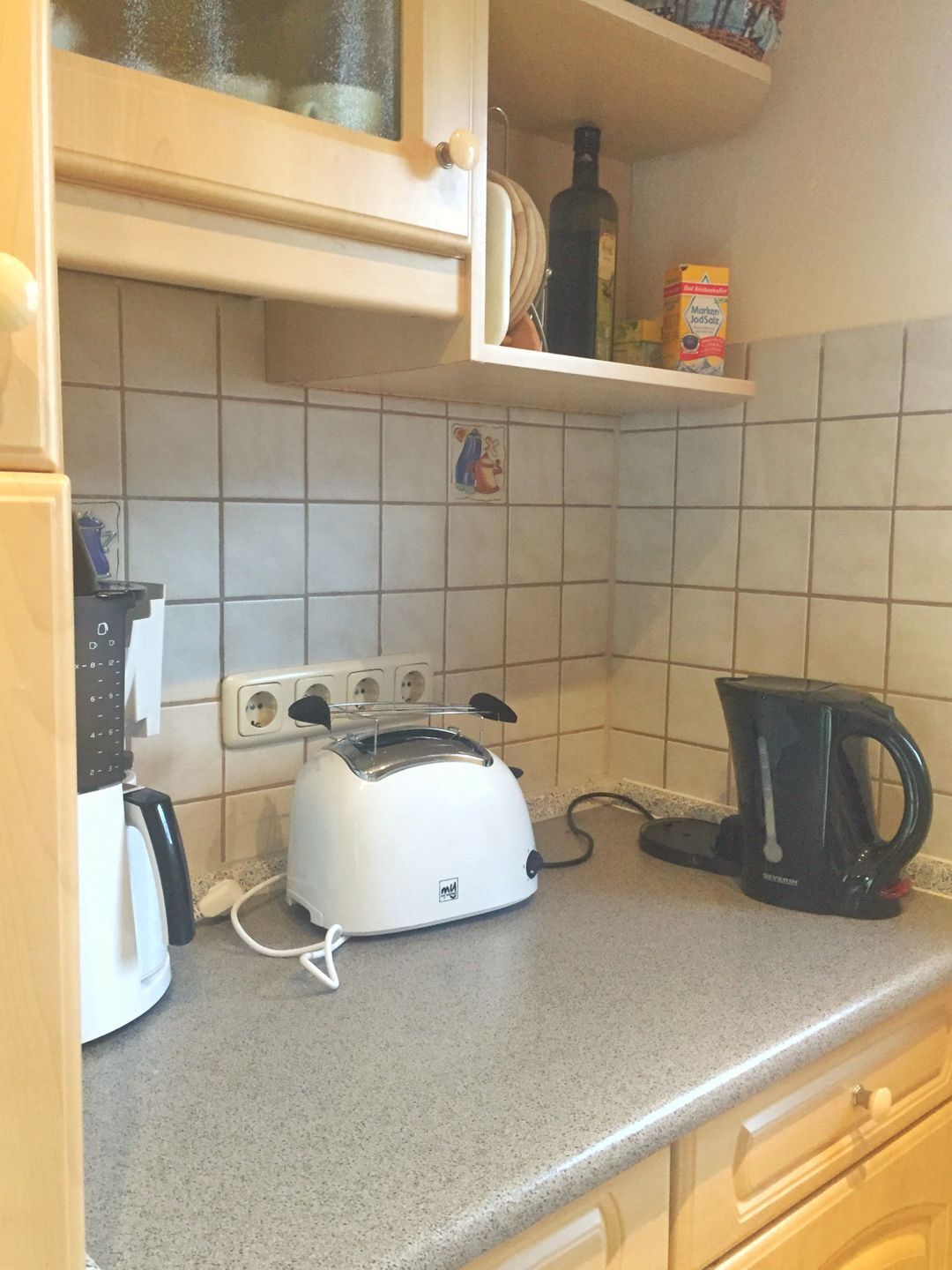 Eine Küche mit einem weißen Toaster, einer schwarzen Kaffeemaschine und einem Regal mit einer Flasche und einer Kiste.
