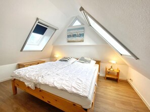 Ein Schlafzimmer mit schräg verlaufender Decke, einem Oberlicht, einem Bett mit weißer Bettwäsche, zwei Nachttischen mit Lampen und einem Gemälde über dem Bett.