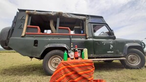 A green off-road vehicle with a covered cargo area, equipped with a spare tire and a table with bottles and glasses. - Amanya Sunset Star Bed Masai Mara (Maasai Mara)