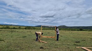 Deux personnes construisent une structure en bois dans un champ ouvert, avec une chaîne de montagnes en arrière-plan.