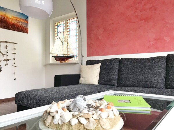 A modern living room with a glass coffee table, a sofa, a red accent wall, a stained glass window, and a painting of a sunset.