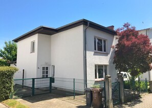 A two-story building with a white exterior, a green metal fence, and a tree with red leaves.