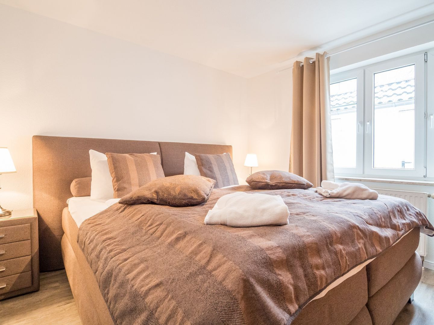 Un lit bien fait, avec une tête de lit marron, plusieurs oreillers et une table de chevet surmontée d’une lampe.