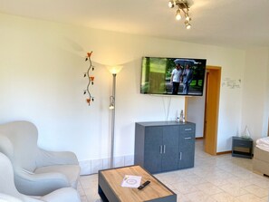 A modern living room with a flat-screen TV, a sofa, a cabinet, a floor lamp, and a small bedside table.