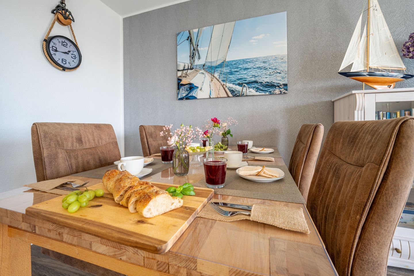 A dining area with a wooden table, chairs, a sailboat painting, and a wall clock.