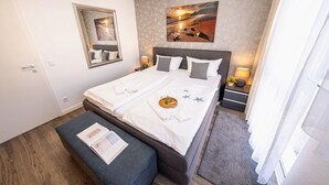 A neatly made bed with pillows, a bedside table with a lamp, a wall-mounted mirror, and a painting above the bed.