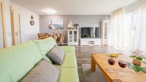 A living room with a green sofa, a wooden coffee table with wine glasses, a TV, and a dining area with chairs and a table.