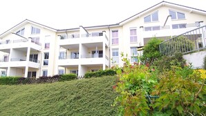A multi-story residential building with balconies, a well-maintained hedge, and a staircase leading to an upper level.