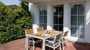 An outdoor patio with a table set for two, surrounded by white chairs, and a large window with a view of the garden.