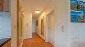 A hallway with tiled flooring, white walls, and a ceiling light. There are doors on the right and a kitchen area on the left.