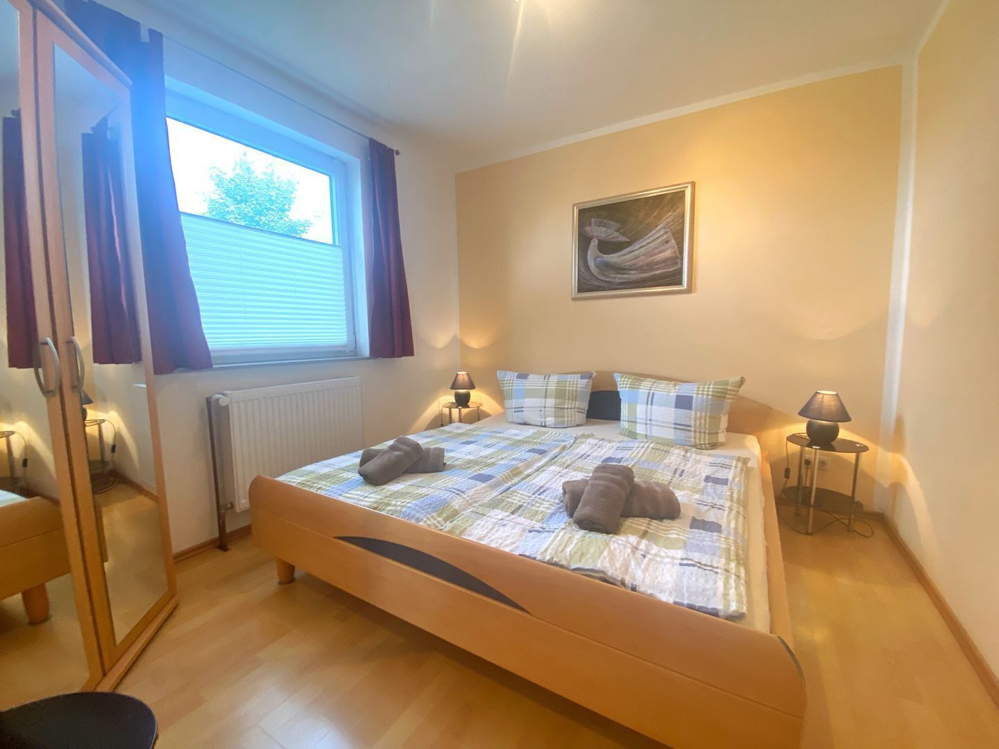 Un dormitorio con cama, dos lámparas de noche, una ventana con cortinas, un radiador, un cuadro en la pared y suelo de madera.