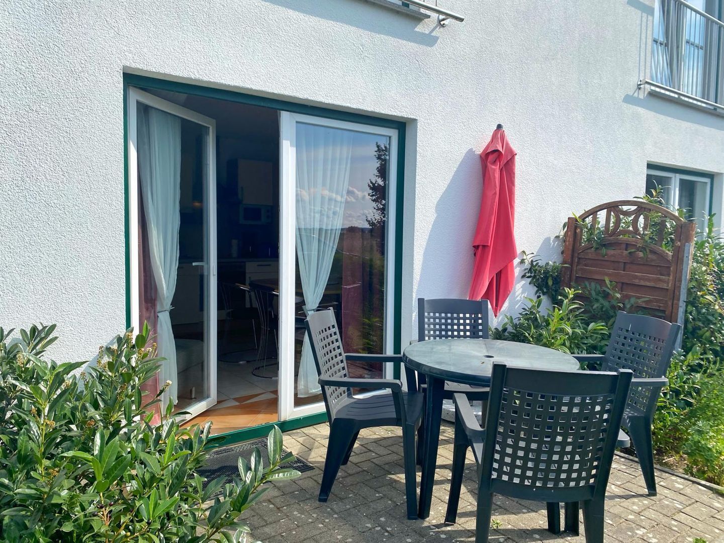 Un patio al aire libre con una mesa y sillas, una puerta corrediza de vidrio que da a una cocina, y un paraguas rojo colgado en la pared.
