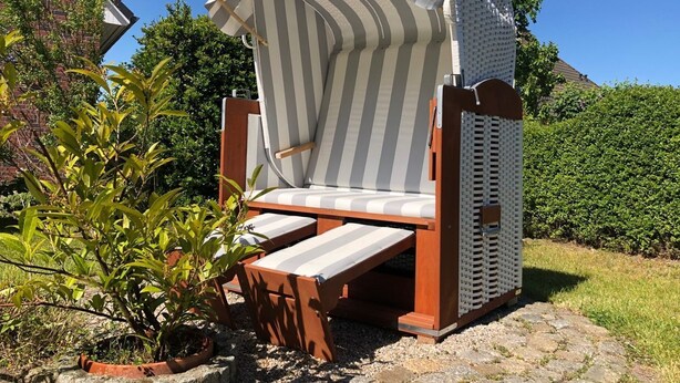 A wooden beach chair with a striped cushion and a white canopy, set on a gravel path with greenery around.
