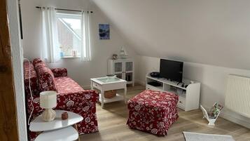 A cozy loft-style living room with a red floral sofa, a white coffee table, a small white cabinet, and a television on a white stand.