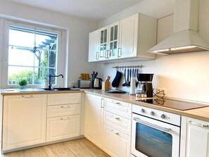 A modern kitchen with white cabinetry, a built-in oven, a coffee maker, and a window providing natural light. - Reethaus Vilmnitz - Ferienwohnung "muglitz" - Reethaus Vilmnitz (Putbus)