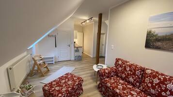 A cozy living room with a red floral sofa, a wooden bench, and a small table. A white door leads to a kitchen and a hallway.