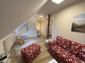 A cozy living room with a red floral sofa, a wooden bench, and a small table. A white door leads to a kitchen and a hallway.