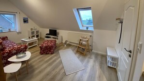 Een gezellige zolderkamer met schuine wanden, een klein raam, een radiator, een witte deur en een knus zitgedeelte met een rode bloemenbank en een kleine tafel.
