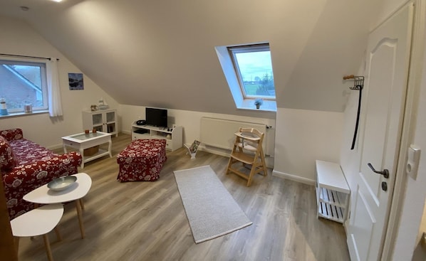A cozy attic room with a sloped ceiling, a small window, a radiator, a white door, and a cozy seating area with a red floral sofa and a small table.