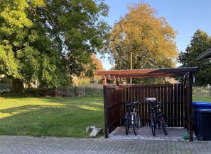 Een overdekte fietsenstalling met een houten hek, een bord dat aangeeft dat deze plek voor fietsers is, en een vuilnisbak.