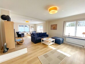 A spacious living area with a sofa, coffee table, and a dining table. There are large windows providing natural light and a view of the outdoors.