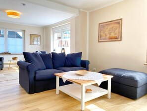 Un salon moderne avec un canapé bleu foncé, une table basse en bois et un pouf assorti. On y trouve une lampe sur une table d’appoint, un tableau encadré au mur et une fenêtre qui laisse entrer la lumière naturelle.