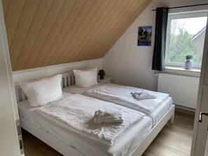 A bedroom with two single beds, a small bedside table with a lamp, a window with curtains, and a radiator.