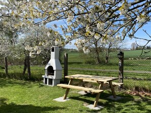 A picnic table with a grill and a chimney structure in a grassy area with blooming trees.