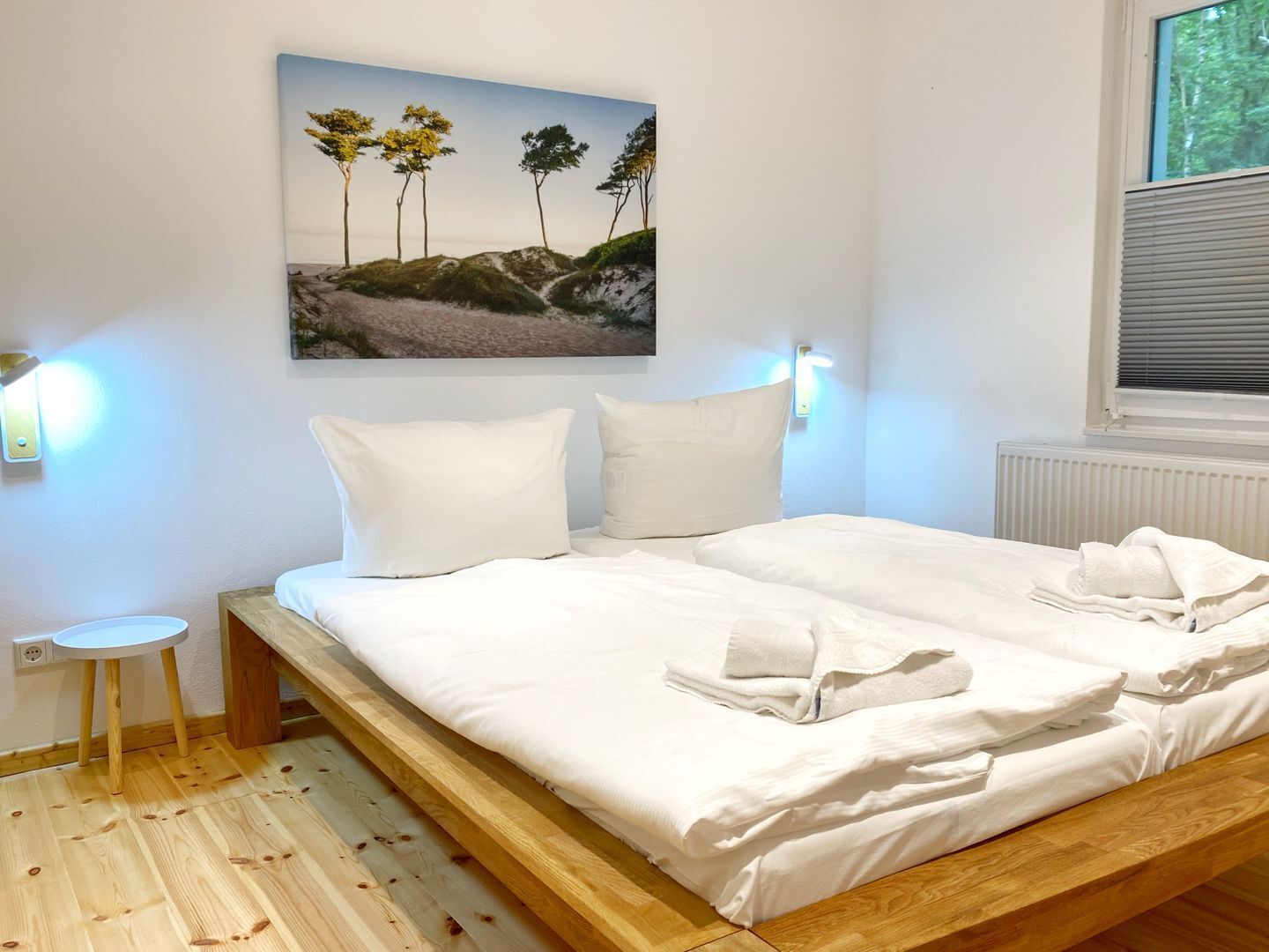 Une chambre à coucher avec un lit en bois, du linge de lit blanc, une petite table de chevet, un tableau accroché au mur et une fenêtre avec des stores.