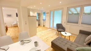 A modern living room with a grey sofa, a dining table with plates and cups, and a kitchen area visible through an open door.