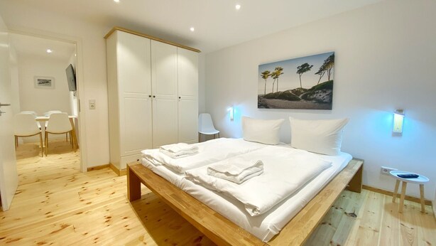 A modern bedroom with a wooden bed, white wardrobe, and a dining area with chairs.