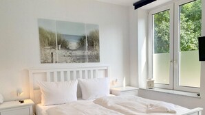 Un dormitorio con una cama blanca, mesitas de noche blancas, una mesita de noche con una lámpara, un cuadro de una playa y una ventana con vistas a la vegetación.
