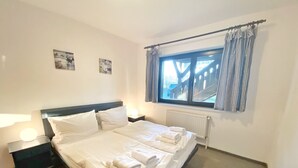 Una cama bien tendida con sábanas y toallas blancas, una mesita de noche con lámpara y una ventana con cortinas.