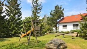 A playground with a wooden swing set and slide, a large rock, and a white building with a red roof in the background.