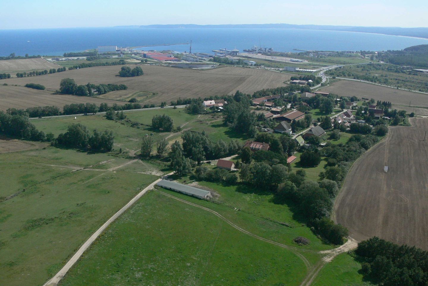 An aerial view of a rural area with fields, a small cluster of houses, and a distant industrial complex.