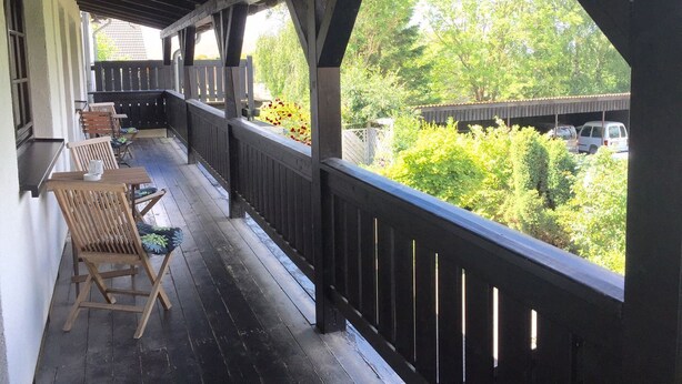 A covered porch with wooden furniture and a view of a parking area and greenery.