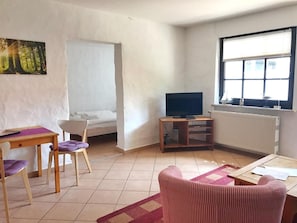 Una habitación pequeña y escasamente amoblada, con una cama, un televisor sobre un soporte de madera, una ventana con persianas y un radiador.