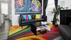 A modern living room with a black leather sofa, colorful cushions, a coffee table with books, a vibrant rug, and a neon cityscape mural on the wall.