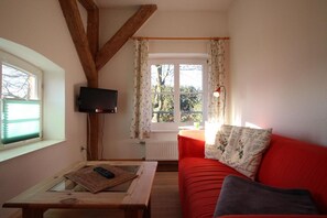 A cozy living room with a red sofa, a glass coffee table with a remote control, a television mounted on the wall, and a window with floral curtains. - Gutshof Dubnitz -Holiday apartment 5 Dörthe Dubnitz near Sassnitz - Gutshof Dubnitz (Sassnitz)