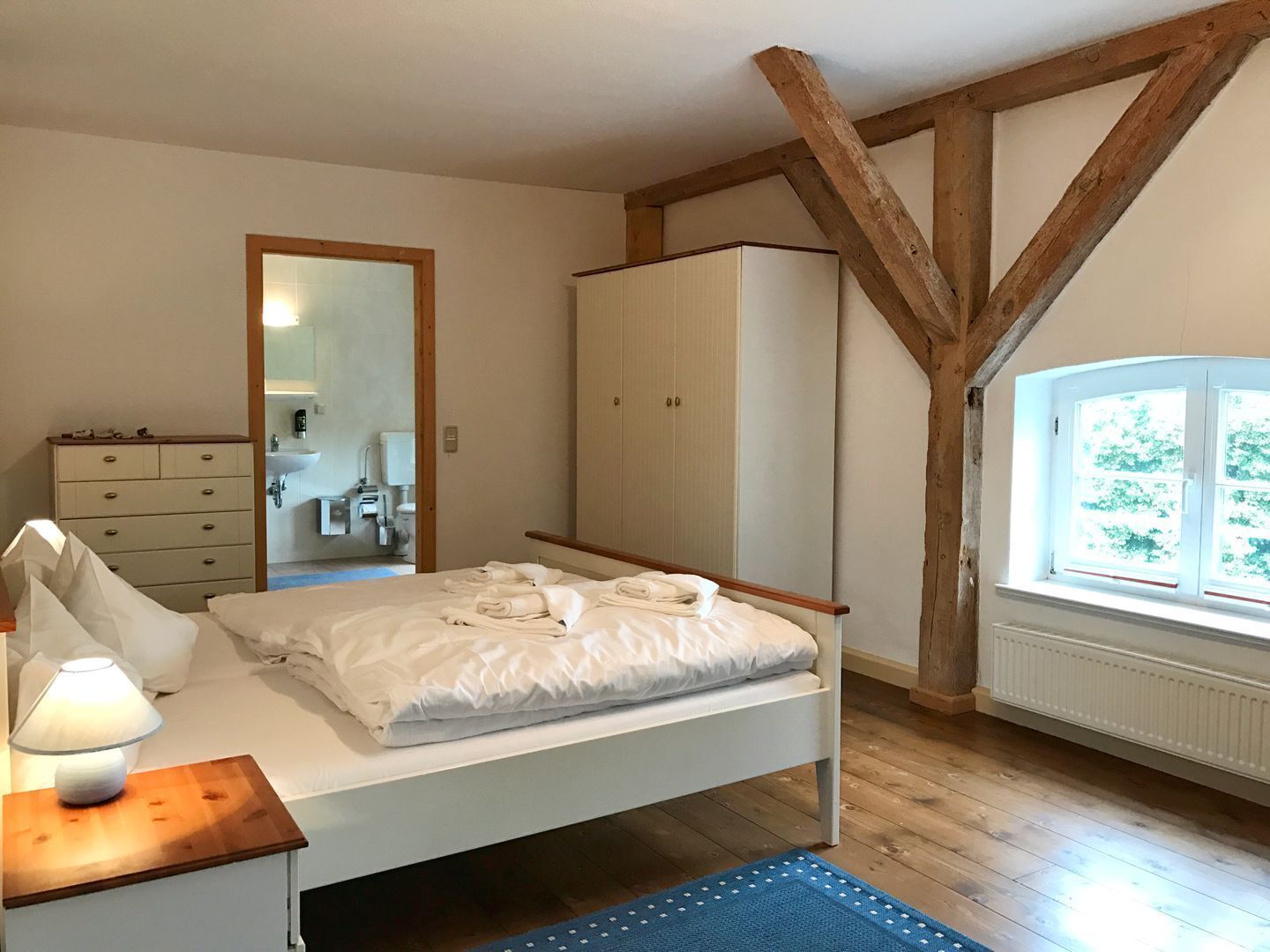 A bedroom with a bed, a dresser, a wardrobe, a mirror, a window, and a lamp.