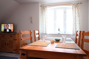 A dining area with a wooden table, four chairs, two glassware items, a green bowl, and a television displaying a person.