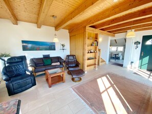 A spacious living area with a wooden ceiling, a sofa, two leather chairs, a coffee table, and a bookshelf.