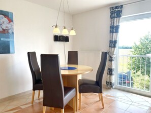 En matplats med ett runt bord, fyra stolar, en blå bordsduk, hängande lampor, en tavla, ett element och en utsikt över grönska genom ett fönster med gardiner.